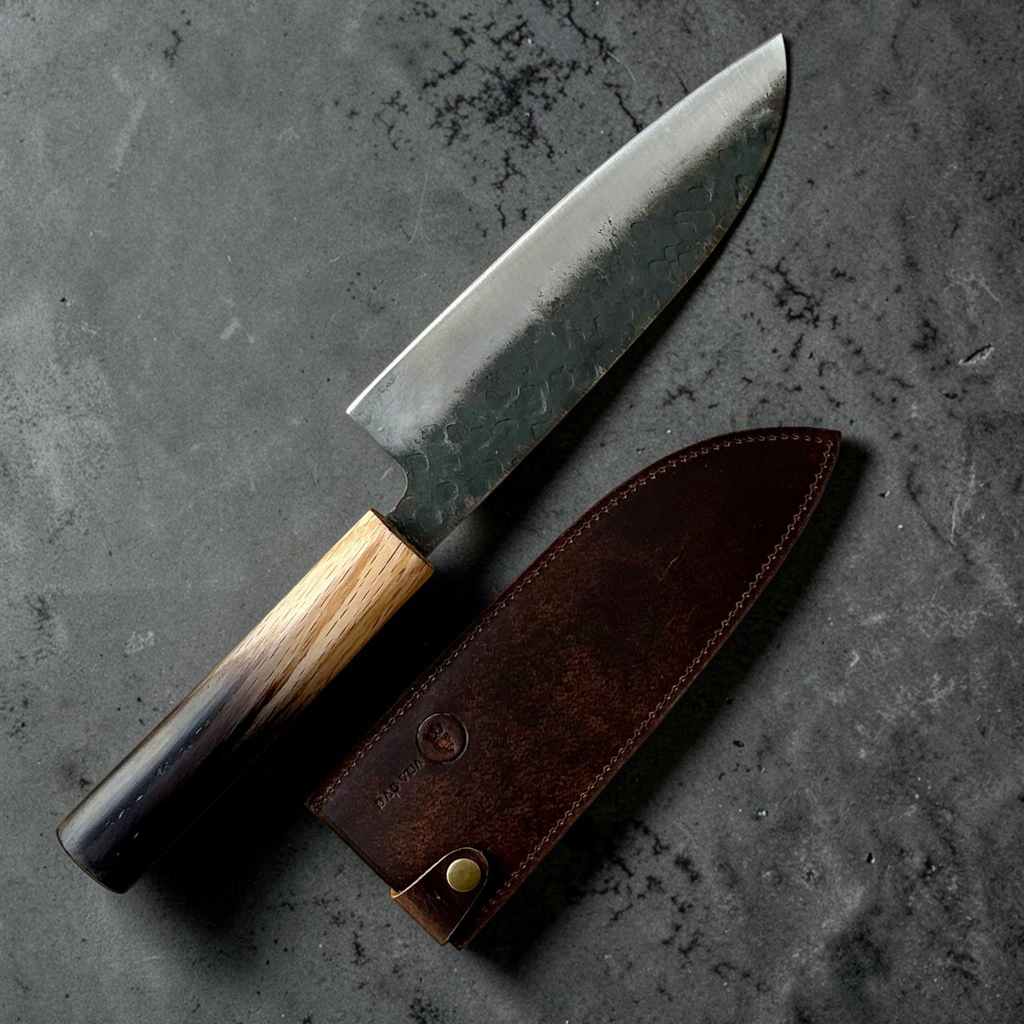 Dao Vua V3 52100 Kuro Tsuchime Santoku beside its fitted leather sheath