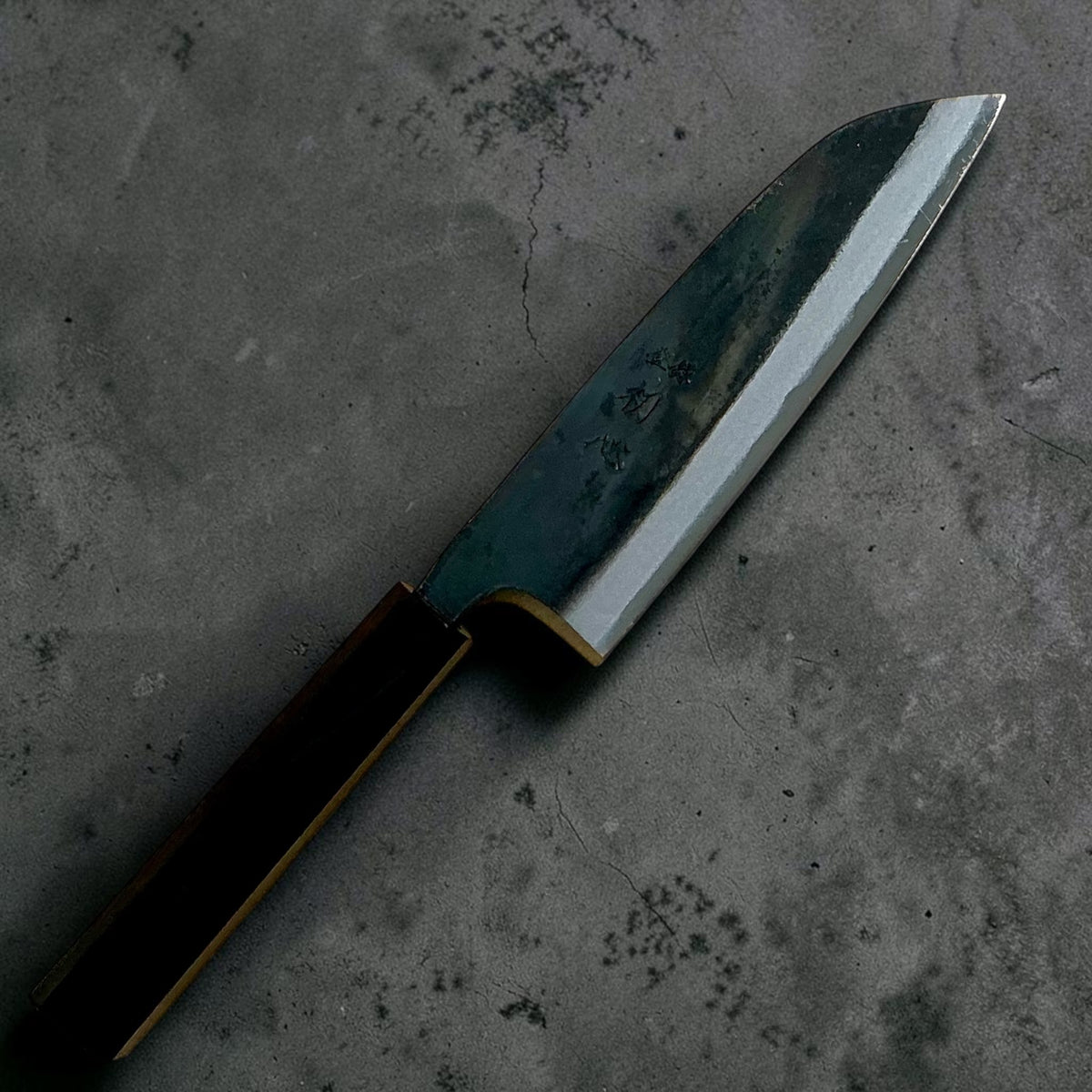 Angled view of the Hatsukokoro Kokugei Blue #1 Santoku showing its kurouchi finish and polished cutting edge.