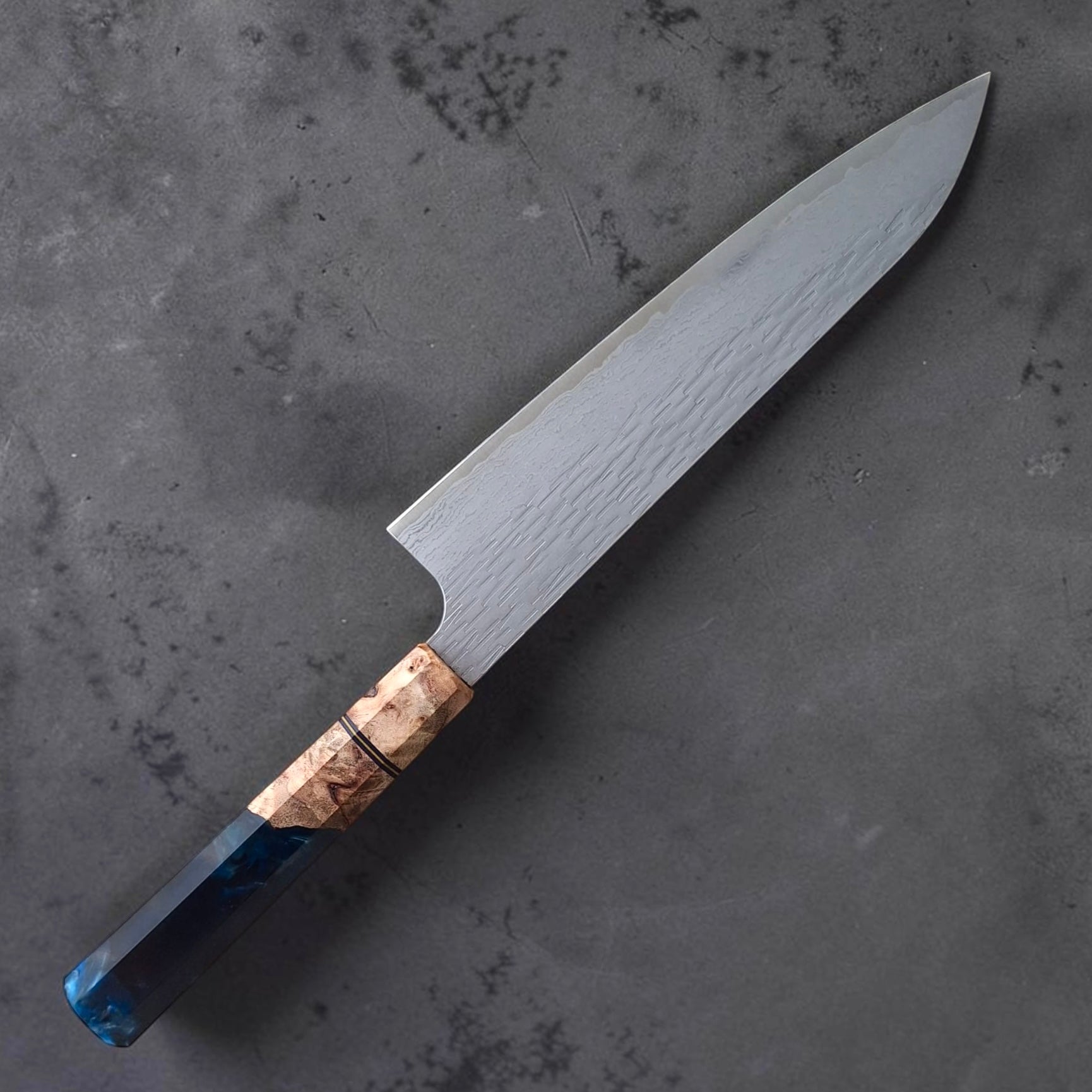 Nigara VG-XEOS Tsuchime Gyuto 210mm with blue Prisma handle, reverse angled full-knife view showing the tsuchime Damascus blade finish on a dark grey surface.