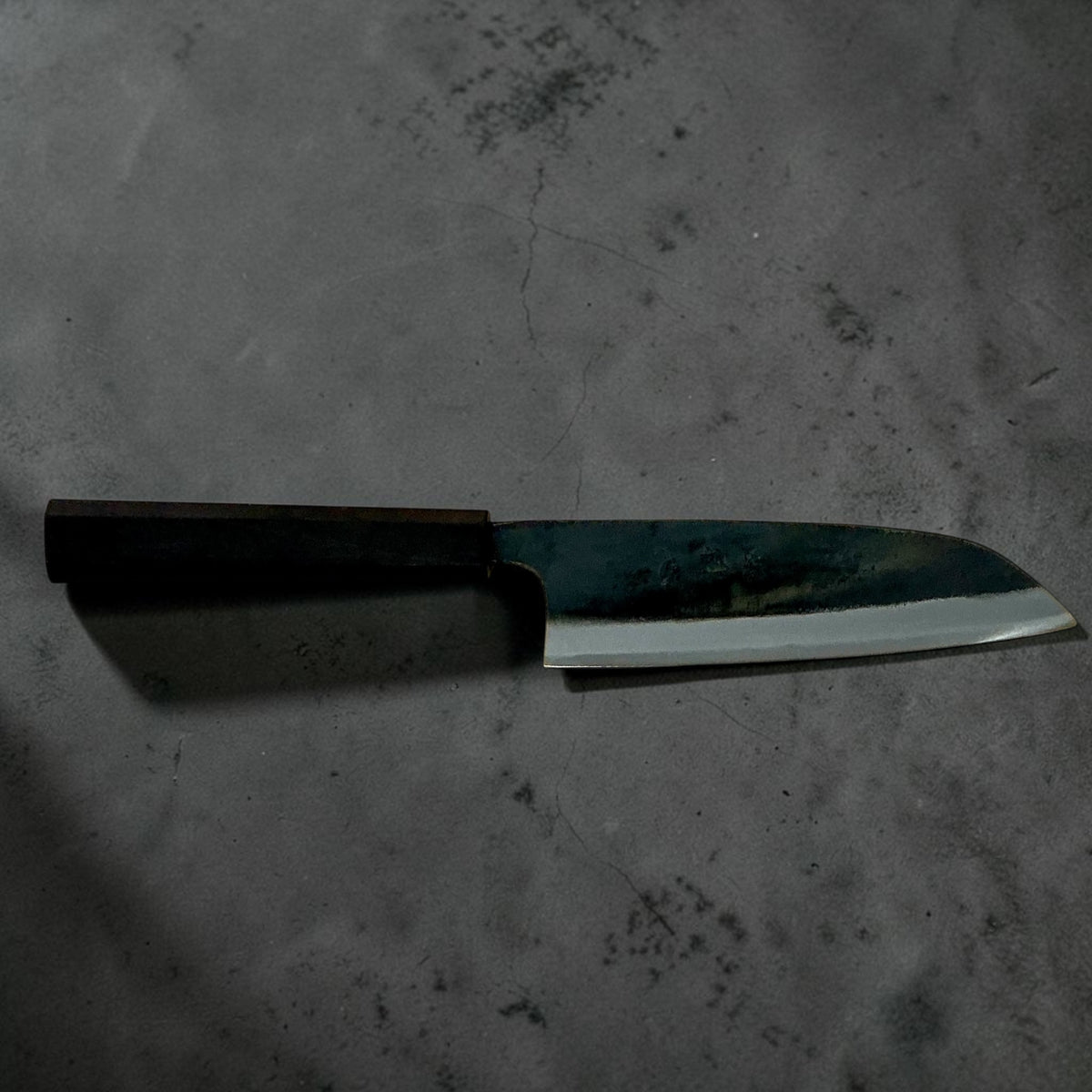 Hatsukokoro Kokugei Blue #1 Santoku 165mm with mono ebony handle, full side profile on a dark textured background.