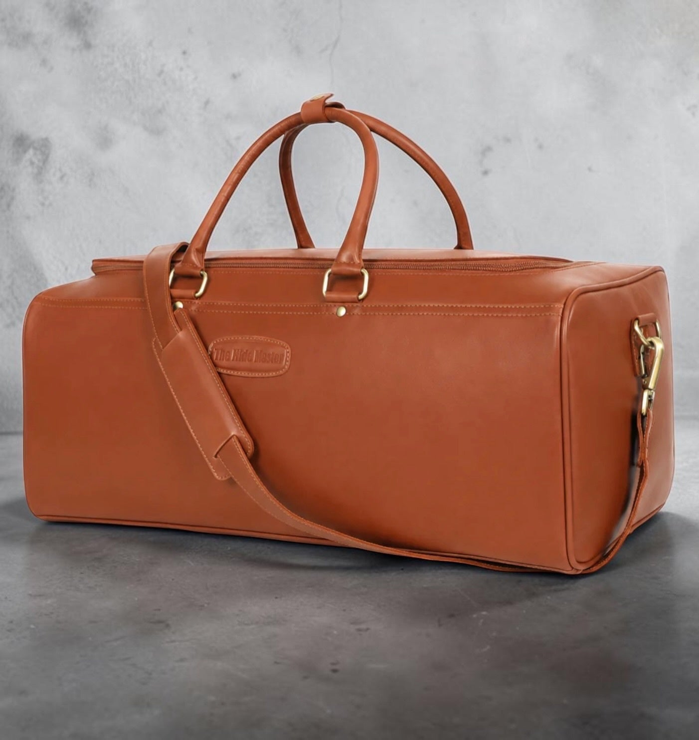 The Hidemaster Aspen leather duffle bag in Cognac, crafted from premium full-grain leather with rolled carry handles, adjustable shoulder strap, and solid brass hardware, shown from a front three-quarter angle.